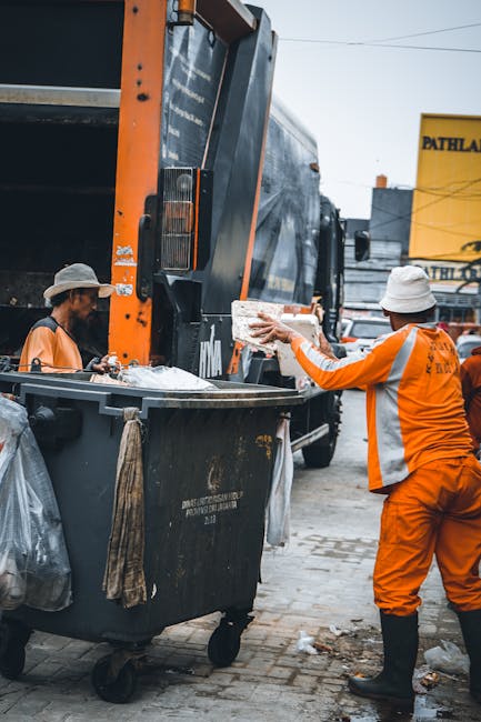 A man wearing a white face mask, light blue shirt, and dark trousers is standing on the back of a large, open-top black industrial waste truck, sorting through miscellaneous household and office furni