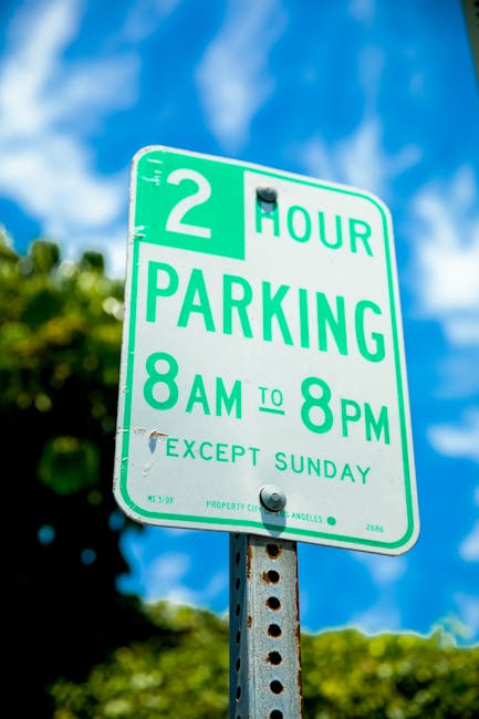 A green and white parking restriction sign attached to a perforated metal pole, displaying a two-hour parking limit from 8 am to 8 pm, excluding Sundays. The sign has bold teal lettering indicating th
