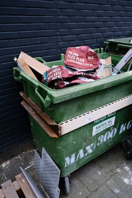 A black wheeled commercial waste container placed on the sidewalk directly in front of a brick-built restaurant with a maroon exterior, large front windows with wooden frames, and signage indicating i