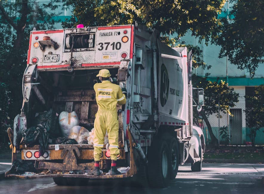 Two workers dressed in bright orange high-visibility uniforms and helmets are engaged in surface cleaning on a paved area, with one holding a long-handled tool and the other standing beside a yellow w
