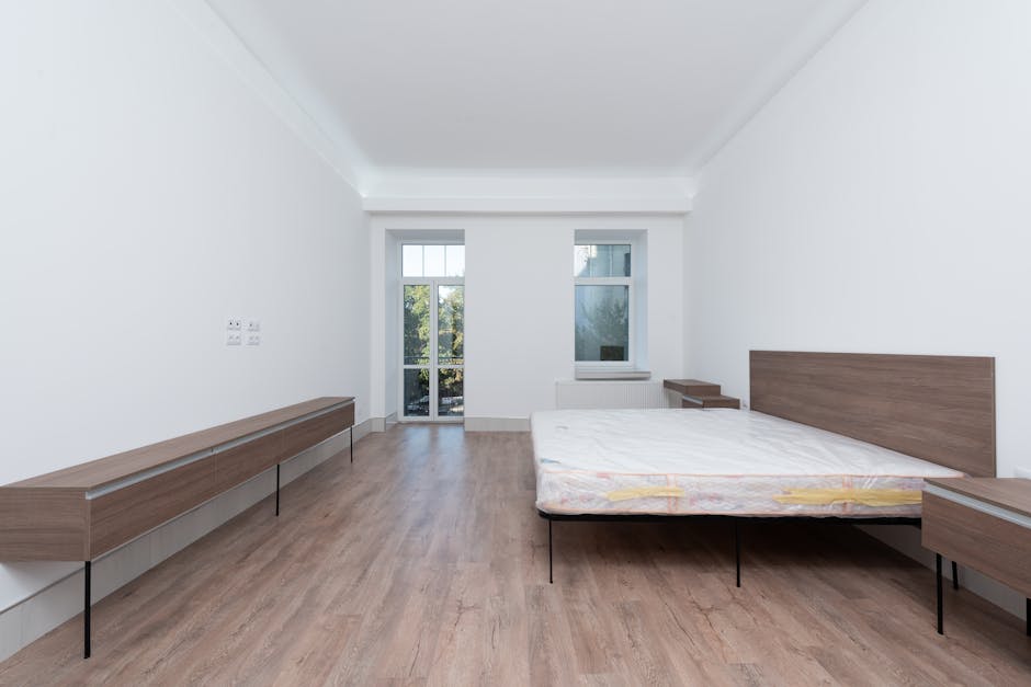 A minimalist bedroom with predominantly white interior design, featuring a plain white wall and ceiling. A white upholstered bed with vertical channel-tufted headboard is positioned in the foreground,