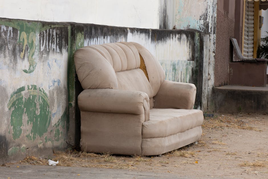 An old, beige upholstered armchair with visible signs of wear and tear, including a torn section on the backrest, is positioned outdoors on a dirt ground next to a weathered concrete wall. The fabric