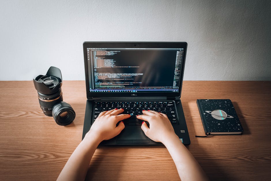 A person's hands are positioned on a black Dell laptop keyboard, with a code editor displayed on the screen. The laptop is placed on a light wooden desk, accompanied by a closed black notebook with a 